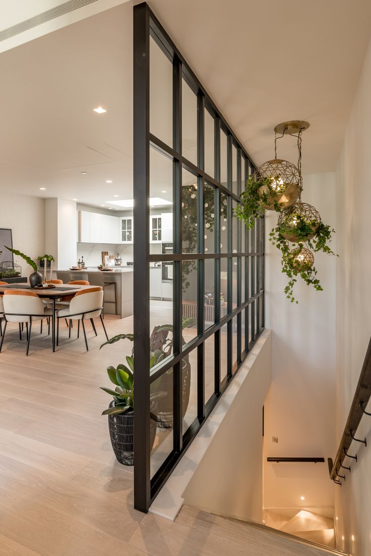 Modern interior featuring a dining area with elegant chairs, a black-framed glass partition, hanging plants with gold accents, and a staircase leading to a bright lower level.